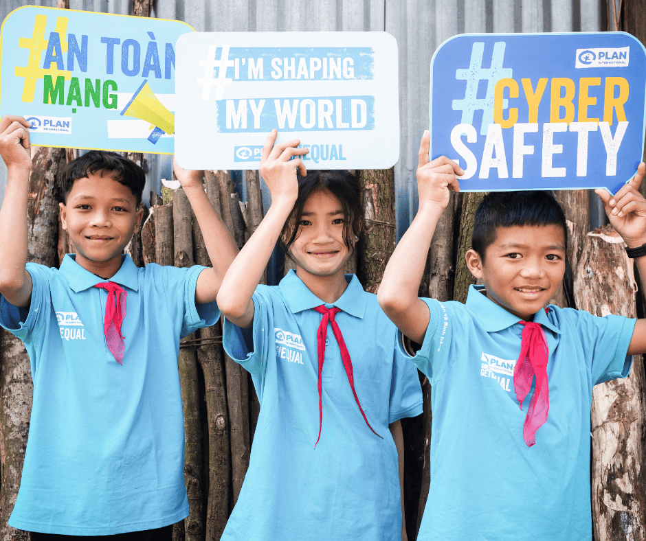 Three young Asian children hold up signs in Vietnamese and English, reading "#I’m shaping my world" and "#Cybersafety" with Plan logos.  The children are wearing their school uniforms, light blue collared shirts with red neckties. 