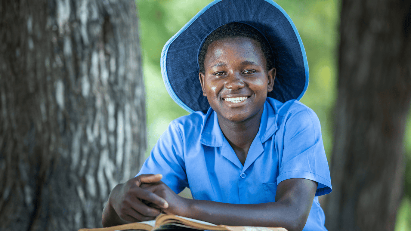 A female high school student from Zimbabwe smiles at the camera. She is sitting at a wooden table outside with a book open, wearing her blue collared school uniform shirt and a wide-brimmed navy-blue hat.