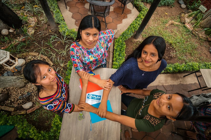 Four Latina young women stand in a circle holding each other’s forearms to form a square between them, which frames the Plan International logo. They all look up at the camera.