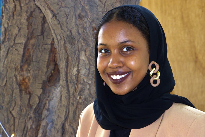 A young African woman stands in front of a tree. She is smiling and wearing a black head scarf secured with a brown and gold clasp.