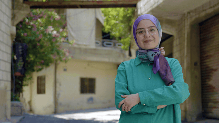 A young woman wearing a head scarf wears a closed-mouth smile as she faces the camera with her arms crossed over her chest. The purple scarf is tied around her neck, and she is wearing a turquoise long-sleeved shirt with silver buttons down the front.