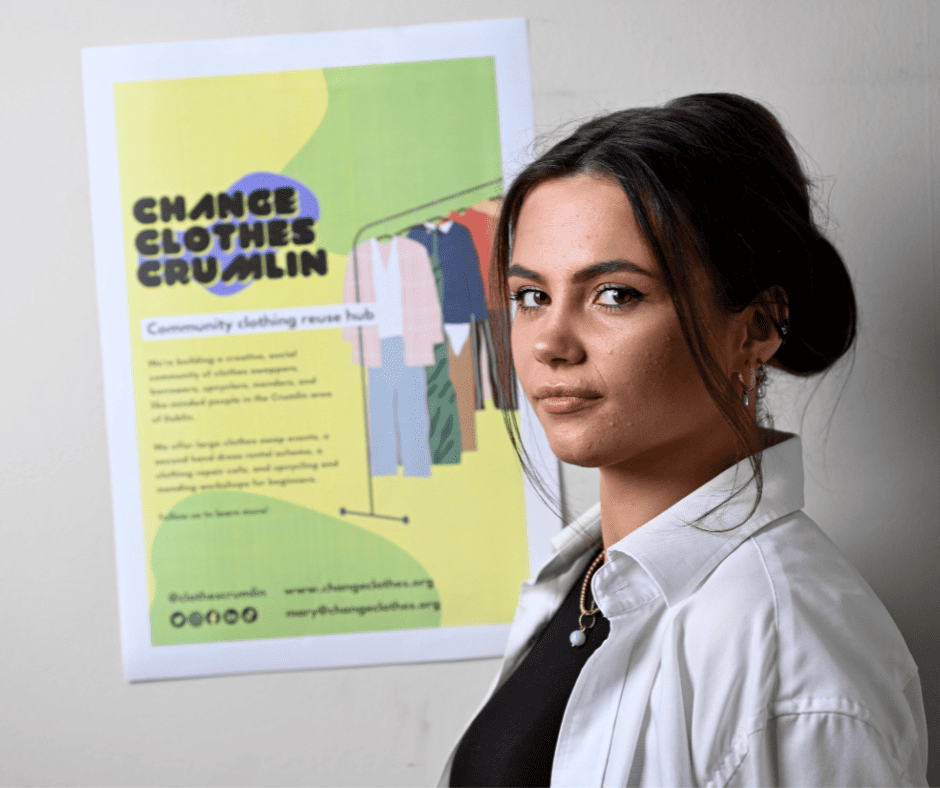 A young woman stands in front of a white wall with a poster reading “Change Clothes Crumlin: Community clothing reuse hub.” Her dark hair is in a bun, and she wears a white long-sleeved collared shirt over a black top.