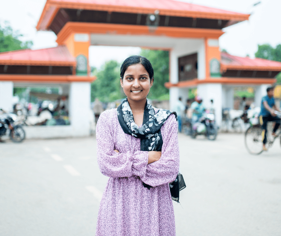 A young South Asian woman smiles and stands with her arms crossed in front of a large gate painted white and orange. She wears a black scarf wrapped loosely around her neck and a long-sleeved dress with small purple dots all over. 