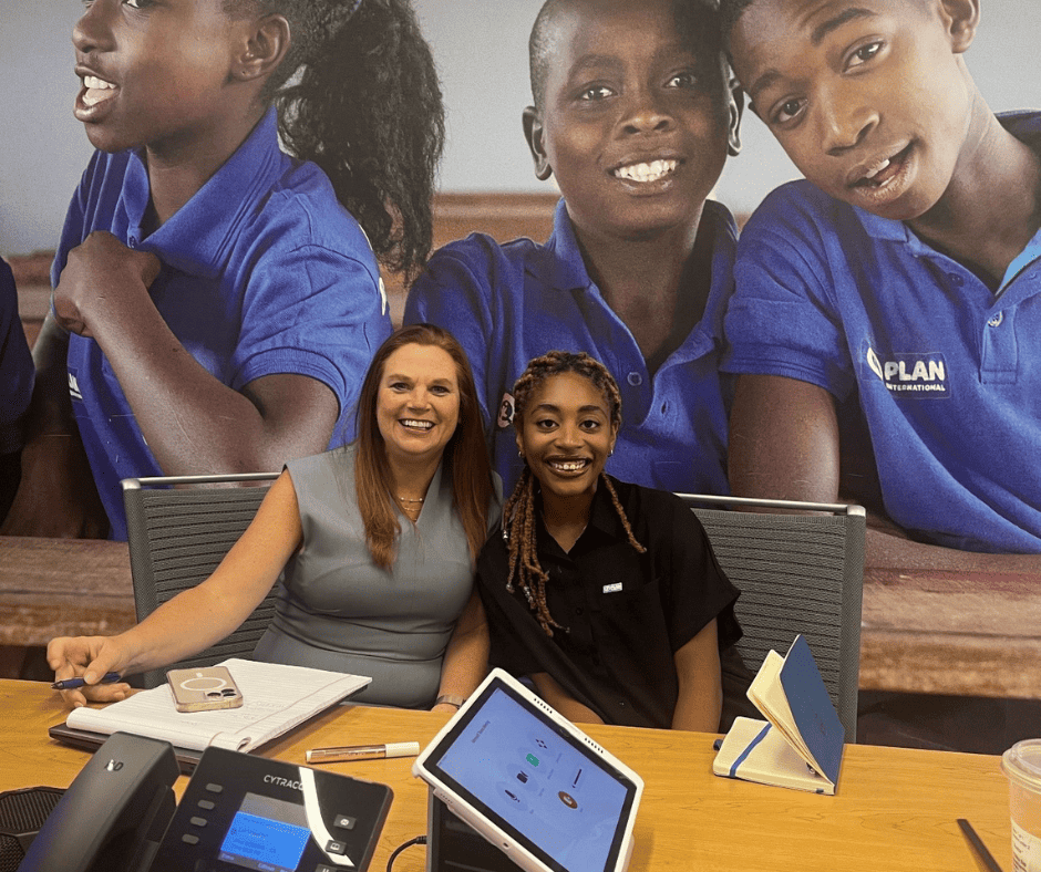 Two women sit side by side in a conference room and smile at the camera.