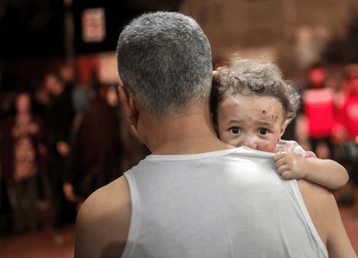 A young child with a frightened look clings to an adult as chaos ensues in the background. Plan International USA responds to assist children during the Gaza-Israel crisis. Donate today.