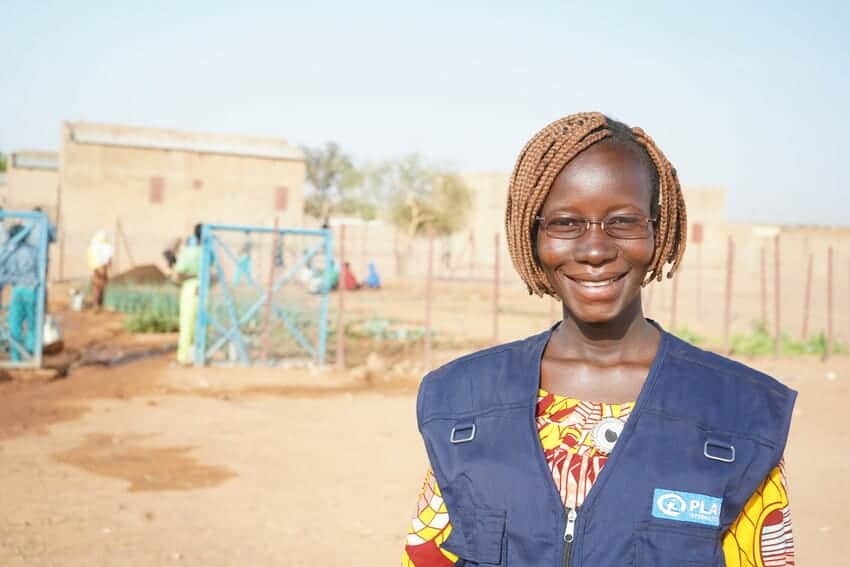 An African woman with short braided hair and glasses smiles at the camera, wearing a dark blue Plan vest over a yellow and red blouse.
