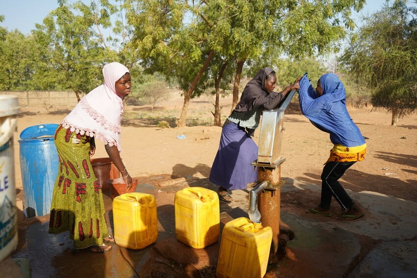 Three girls gather around a water pump, two pushing down on the pump and the third reaching down to grab a large yellow jerry can. 
