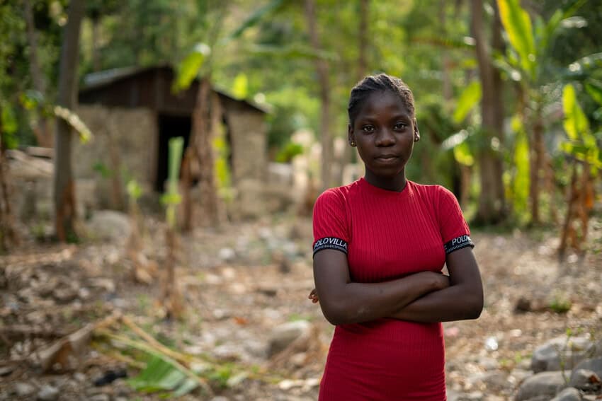 A young Black woman looks at the camera with her arms crossed. She wears a red dress and stands in front of palm trees and a small shed that is blurred in the background.