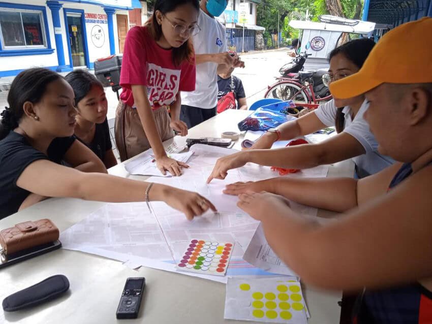 Seven young people sit around a table, some leaning over it to point at maps laid out in front of them. They are outside, and the building behind them has a wall that reads “Community center.”