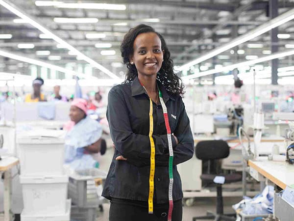 A young women in black clothing stands in front of a large room with people sewing in the background.