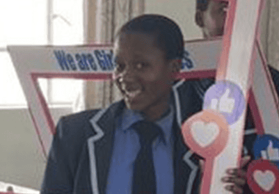 Amanda, a young Zimbabwean woman, smiles for the camera while wearing her school uniform, which includes a navy blue blazer over a light blue collared shirt and a navy blue tie.