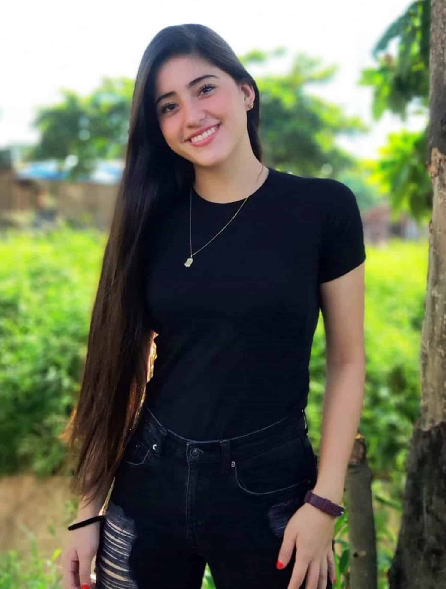 Maria Alejandra, a young Bolivian woman, smiles at the camera, wearing a black t-shirt tucked into black distressed jeans.