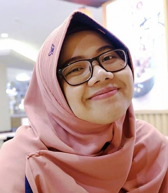 A young woman in Indonesia wearing a pink head scarf and glasses smiles for the camera.