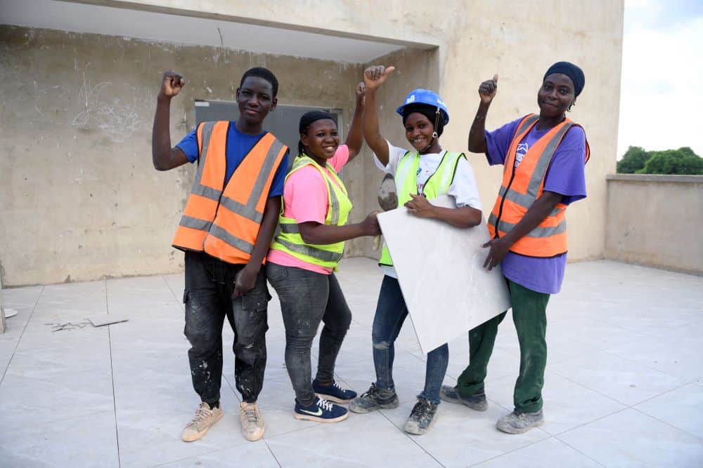 Three women and a man in Ghana stand on an outdoor patio, wearing neon construction vests and smiling as they give the camera a thumbs up.