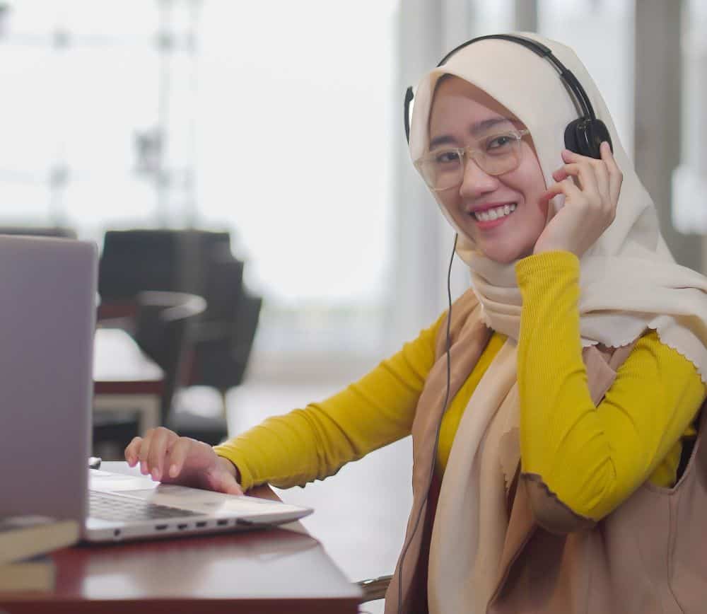 A young woman in Indonesia sits at a table in front of a laptop, wearing a tan headscarf and a yellow shirt with a brown vest. She has a black headset on and is smiling at the camera.