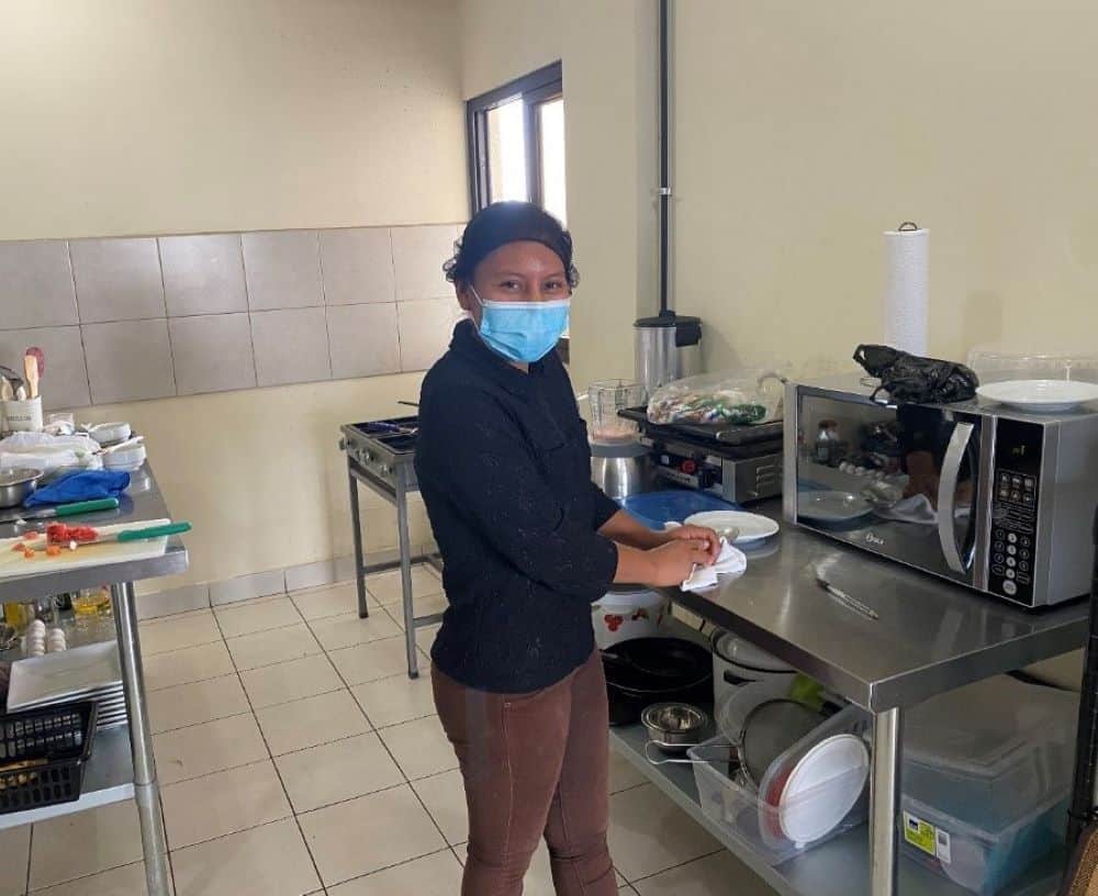 Eliza, a young Guatemalan woman, poses for a picture in a hotel kitchen, standing next to a microwave on a stainless-steel table while wearing a blue surgical mask and black hair net.