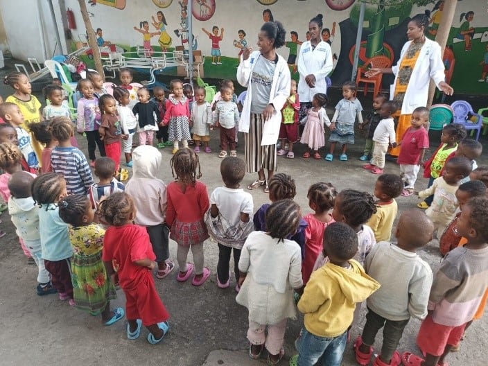 A group of young children stand in a circle in front of a brightly decorated building, gathering around a woman wearing a white coat.