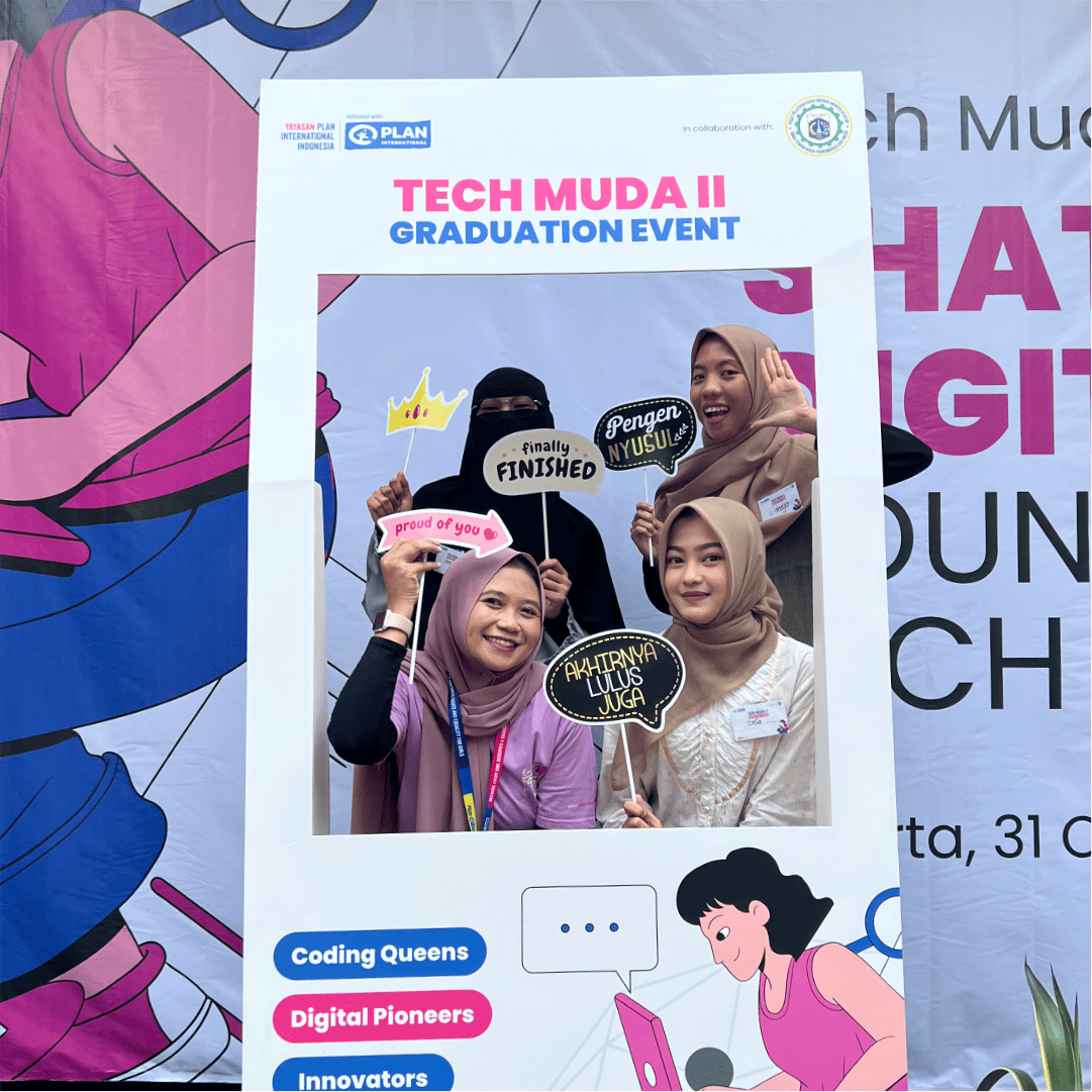 Four women wearing head scarfs pose for a photo behind a cardboard cutout that says “Tech Muda II Graduation Event” at the top and the words: Coding queens, digital pioneers and innovators at the bottom.