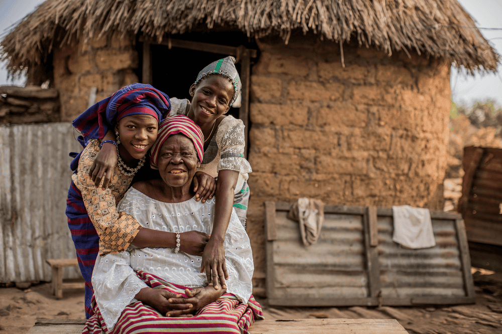 Denise wearing yellow, blue and red stands next to her mother Esther as they both lean over and hug grandmother Inna, who is wearing a white top and r striped skirt.