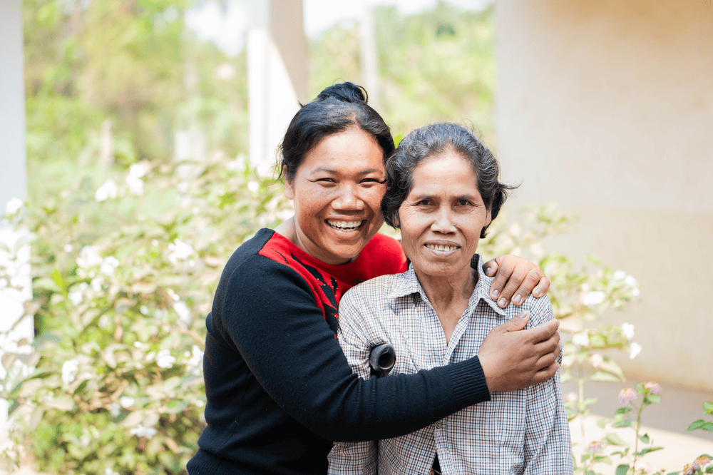 Sokna wearing a blue and red sweater hugs her mother Bui Non as both smile and face the camera.