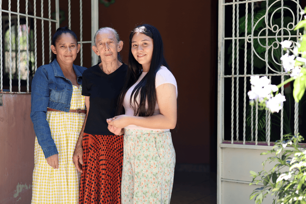 Hazel (right) stands in front her familyās home with her mother Ana (left) and grandmother Paz (center).