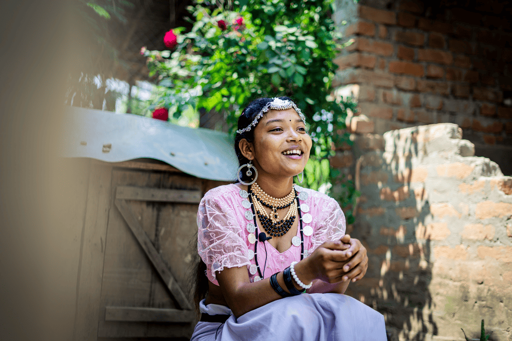 Pushpa sits on steps outside her family's home wearing a pink top and white pants and an assortment of beaded jewelry.