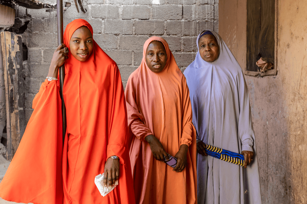 Teenage Rahamatu holds onto a post and stands to the left of her mother and grandmother, all three of them holding menstrual pads.