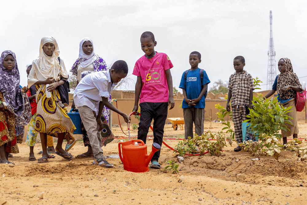 Young children in Niger gather while two boys fill a red watering can in the middle of a sandy school garden.