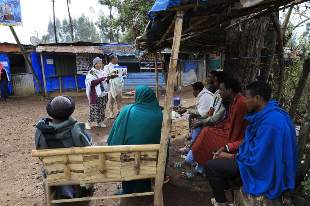 A woman speaks to a group of young men sitting under a wooden overhang in Ethiopia.