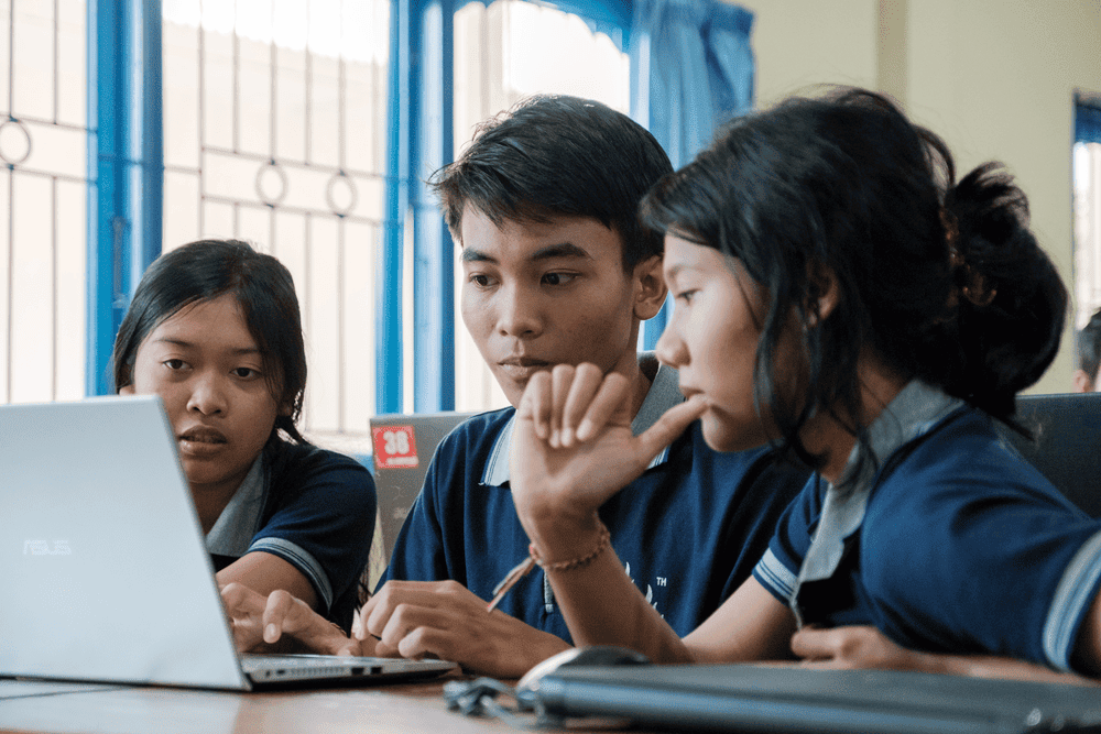 Two girls and one boy wearing blue polo shirts sit in front of a laptop screen.