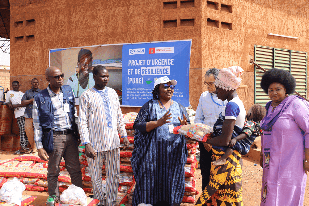 Plan International staff hand a bag of food supplements to a young woman with her child strapped to her back.