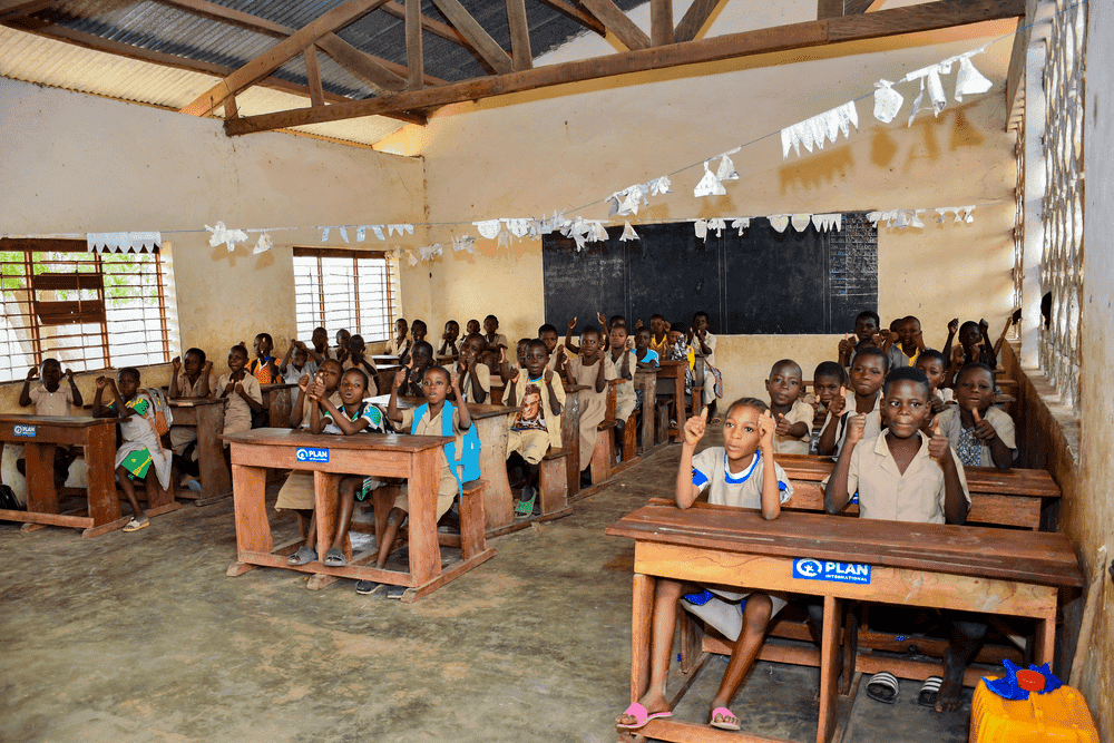 Dozens of children in Benin sit in rows of school desks with Plan International logos attached to the front faces of each desk.