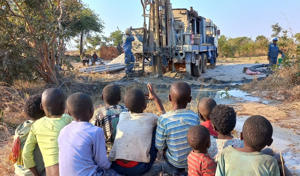 Ten young children in Zambia watch diggers drill a well in their community.