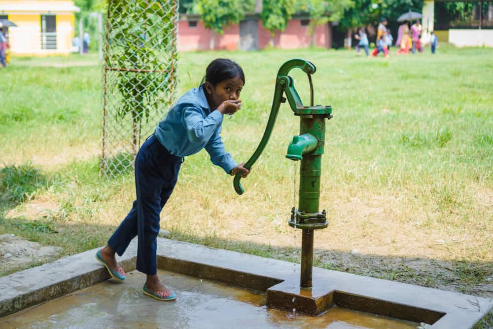 Girl in Nepal wearing a light blue shirt and dark blue pants holds the handle of a water pump as she drinks from her other hand.