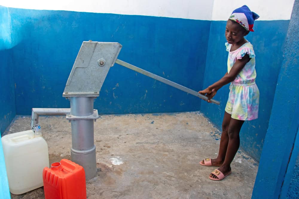 Girl in Haiti holds the handle of a water pump as water fills a white plastic jug. 