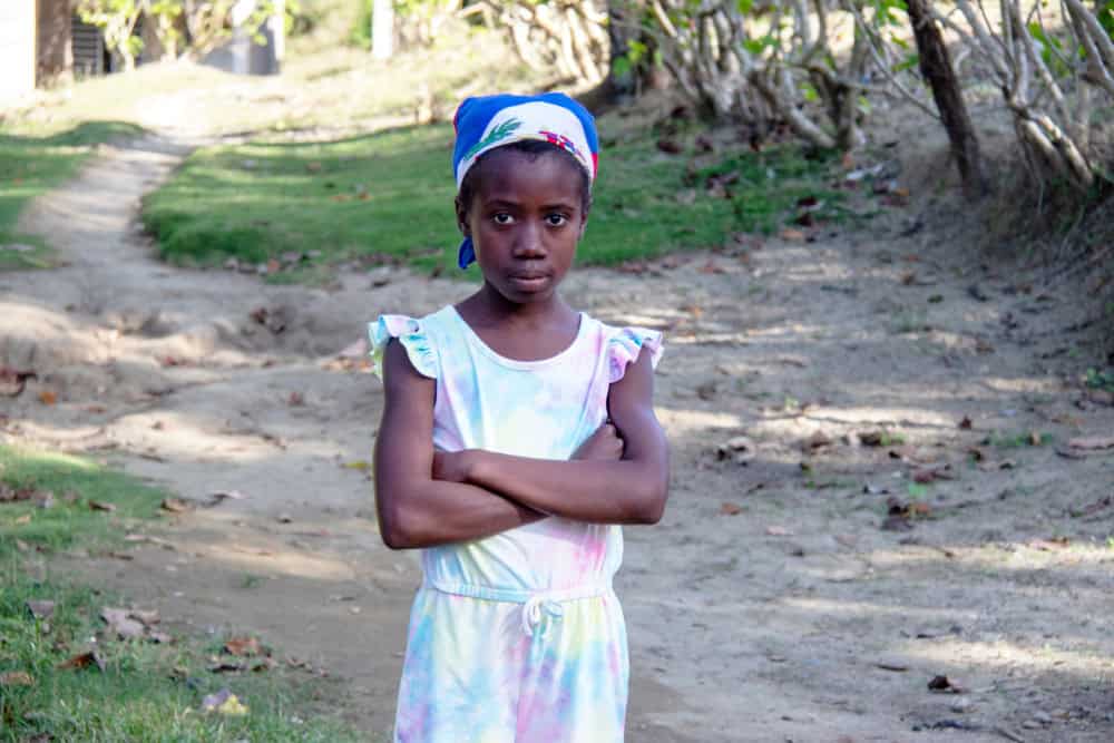 Semi-wide shot of a girl in Haiti standing with her arms crossed as she looks into the camera with a serious facial expression.