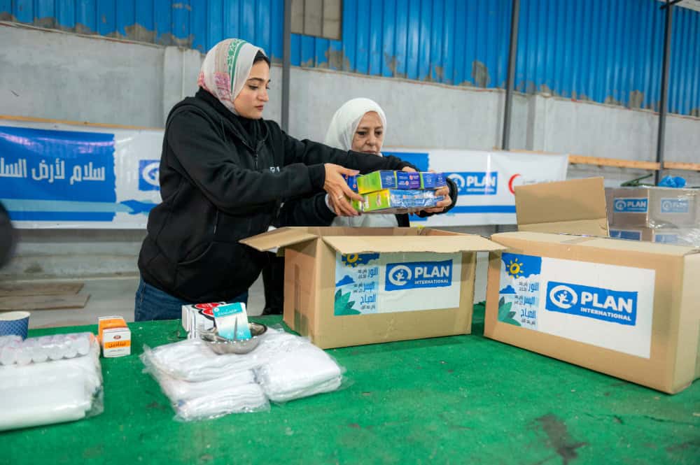 Women pack small colorful boxes into larger cardboard boxes with the Plan International logo on the outside.