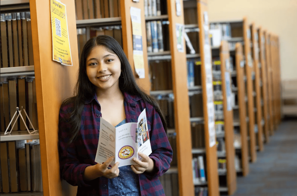 Himangini stands in a library at the end of a bookcase holding a Geaux Girls STEM brochure and smiles at the camera.