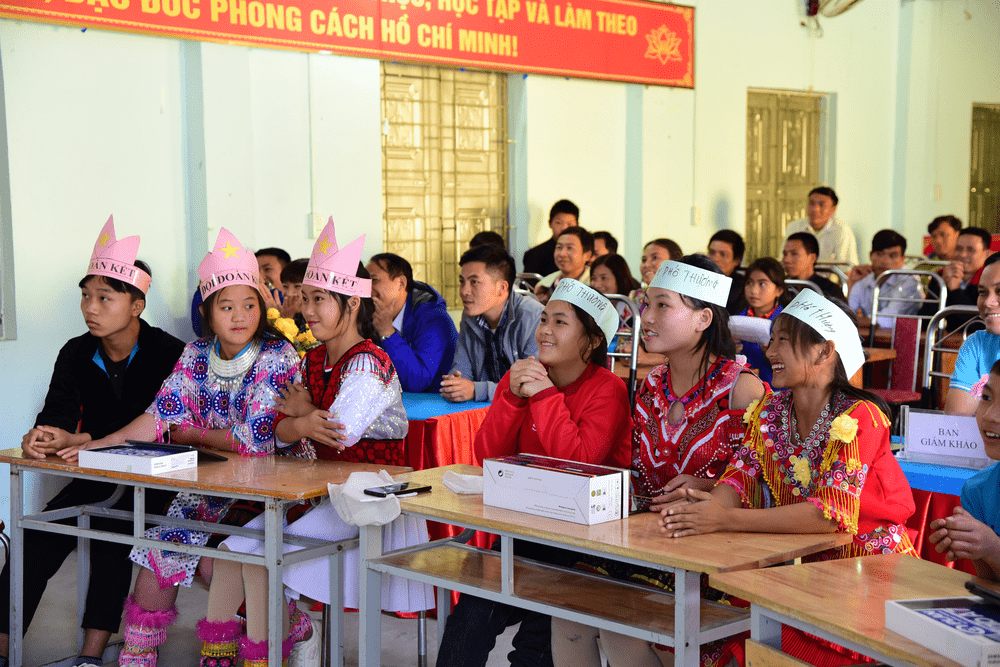 A row of young people with paper crowns sit at desks in front of a classroom full of students.