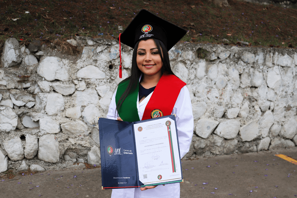 Photo of a young woman wearing a white graduation gown with red and green sashes and a black cap holding open her university diploma.