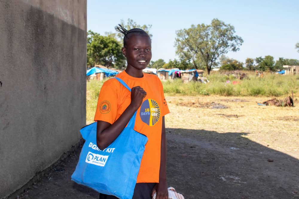 Young woman in South Sudan wearing a bright orange t-shirt holds a blue tote bag with the Plan International logo and smiles.