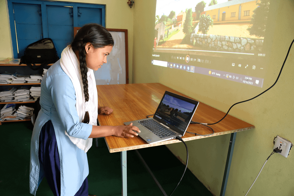 Girl scrolls on computer at desk while screen projects onto wall.