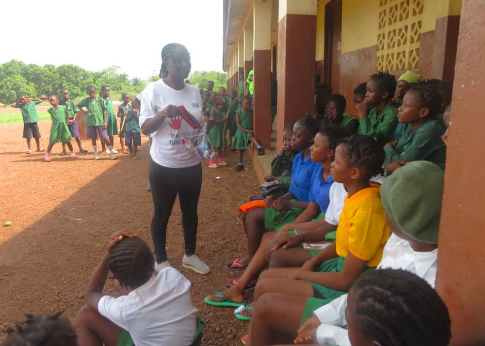 A woman in a white t-shirt stands in front of a group of girls sitting along a wall as more children in green stand in the background.