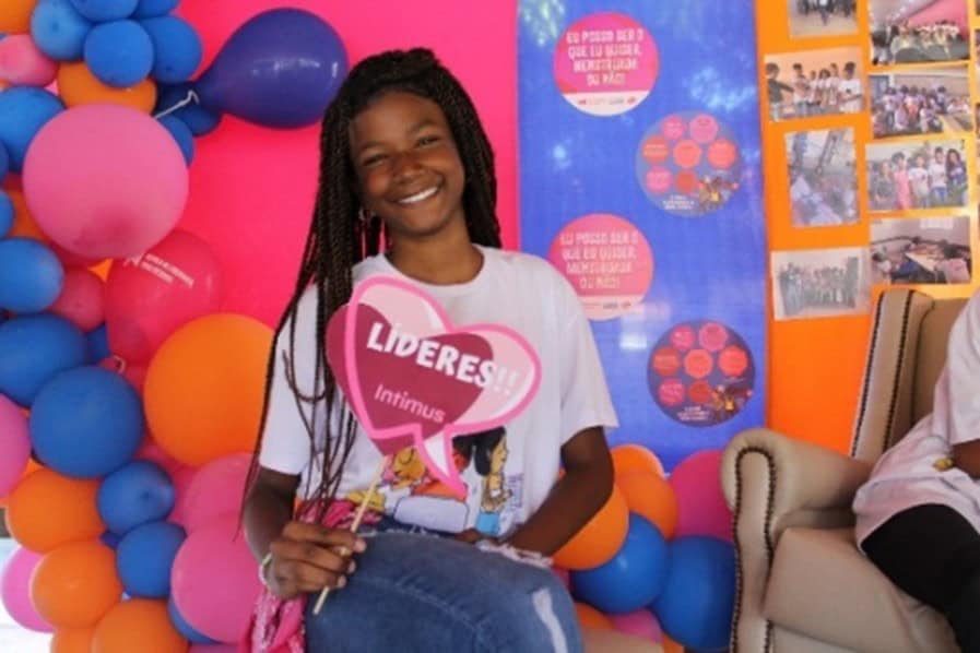 A girl sits in front of balloons holding a heart shaped sign and smiling for the camera.