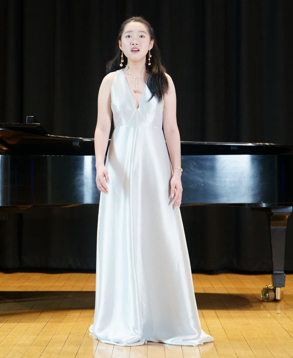 Sophia stands in front of the piano and sings at her recital.