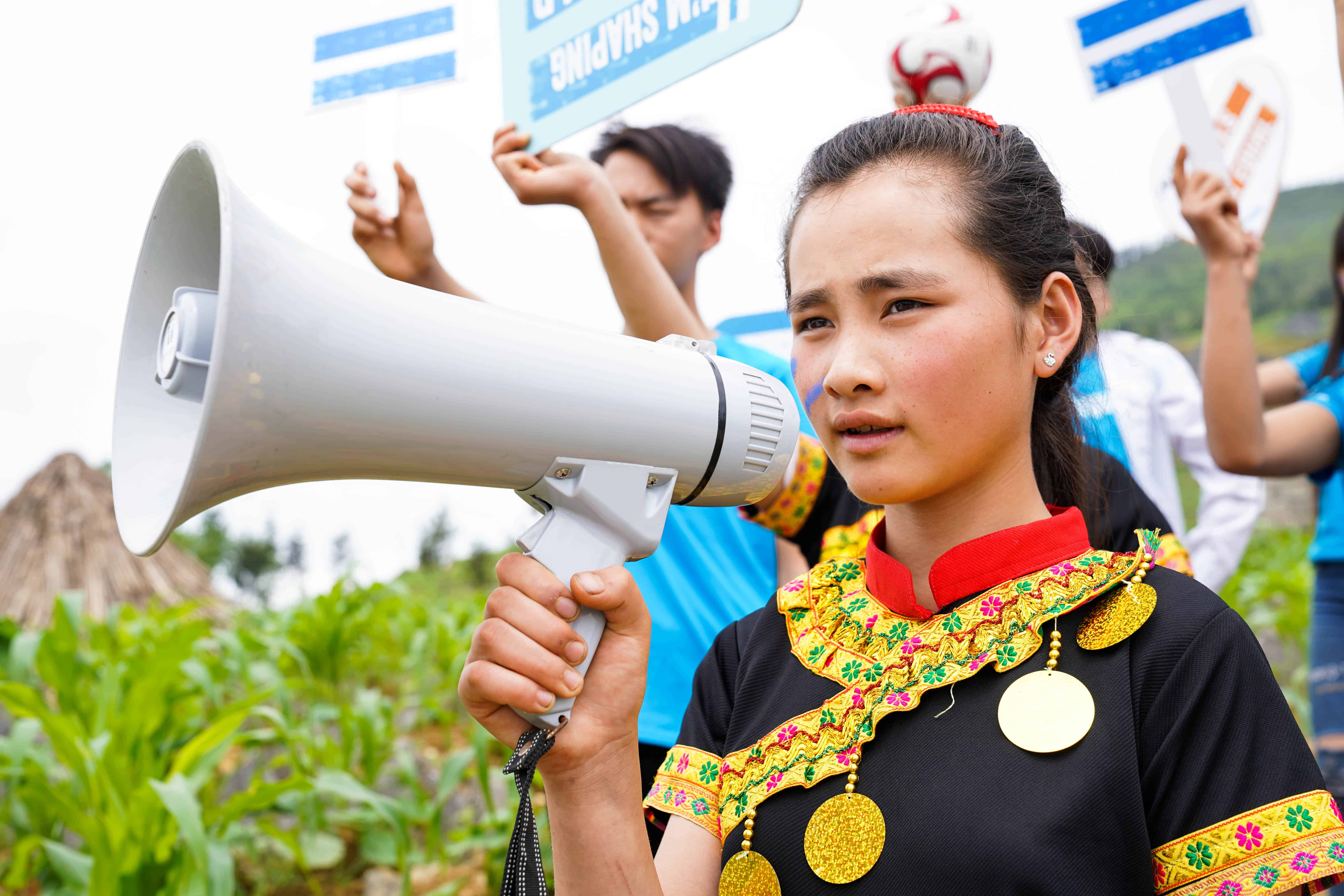 Close-up of a young activist in traditional clothing holding a megaphone, with other youth holding signs in the background, promoting gender equality in Vietnam.