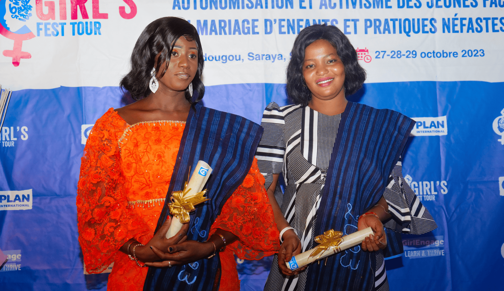 Two young women dressed in traditional Senegalese clothing hold certificates and smile at a high school graduation ceremony in Kédougou, Senegal, celebrating their achievements as part of Plan International's Girls Learn & Thrive project.