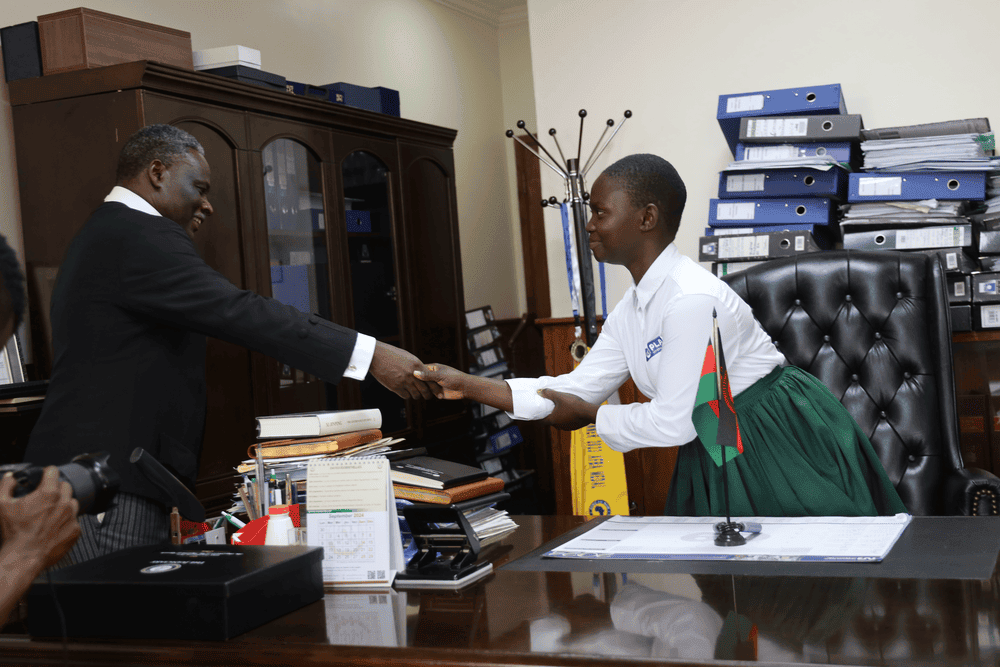 A girl wearing a white shirt and green skirt and shakes the hand of Chief Justice Rezine Mzikamanda.