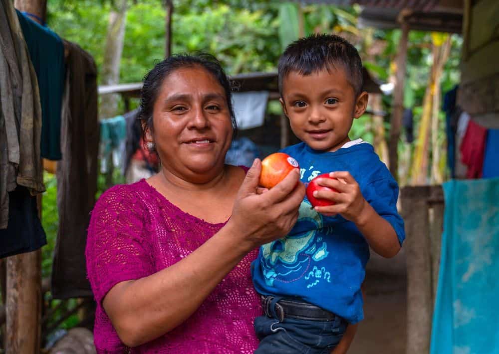 A mother holds her son in her arms as they both hold up apples and smile for the camera.