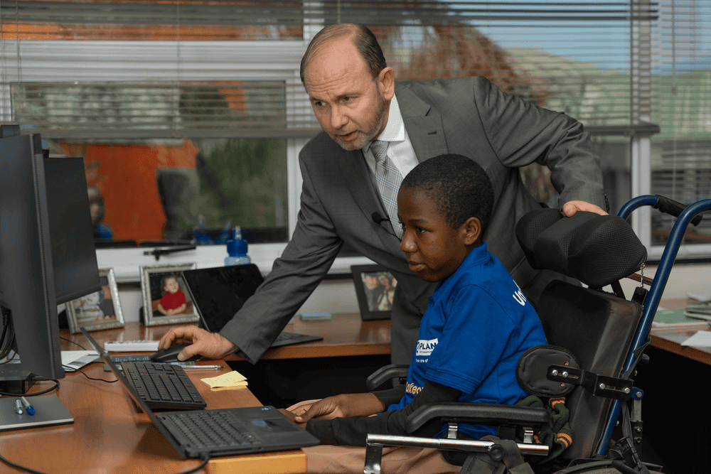 Peace sits in a wheelchair at a desk as World Bank Malawi country manager leans over and directs her on a computer.
